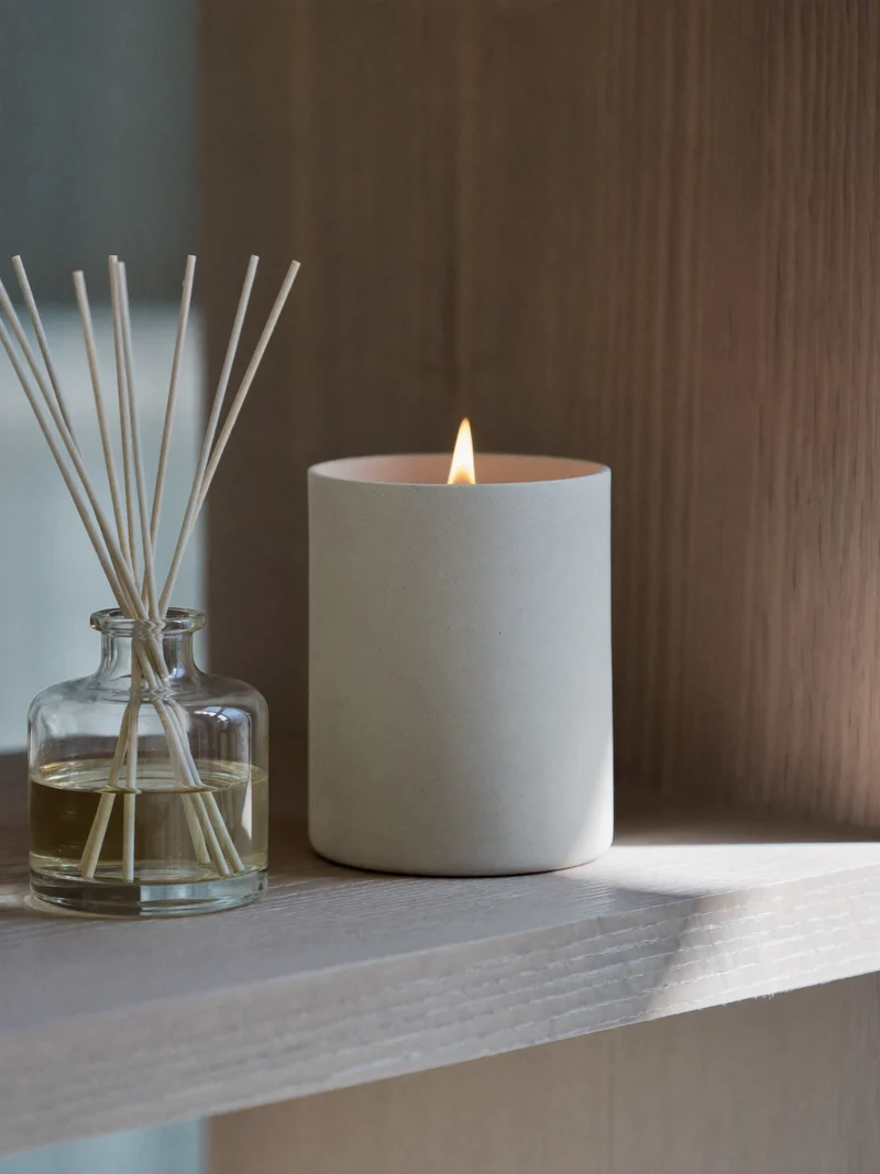 Keramisk duftlys med tændt væge og slank rørdiffuser på en træhylde